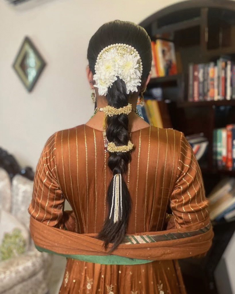 Elegant braided hairstyle decorated with white chrysanthemums, pearls, and gold accessories, paired with a rust-brown embroidered Punjabi outfit.